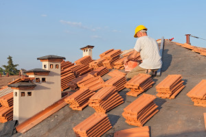 roofing: construction worker on a roof covering it with tiles - roof renovation: installation of tar paper, new tiles and chimney