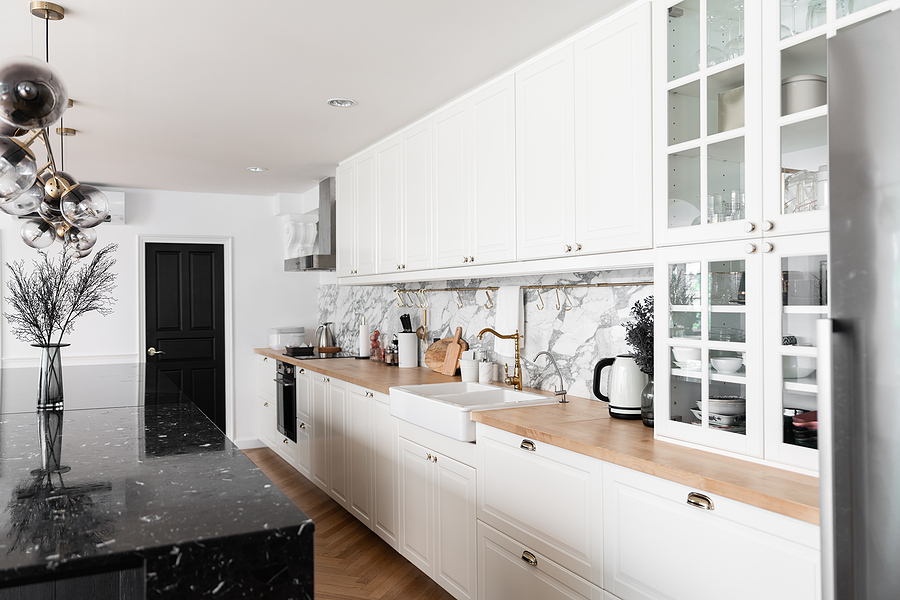 Modern classic kitchen interior with kitchen appliances and white ceramic sink with gold mirror faucet on wood top with marble wall in the background / kitchen interior