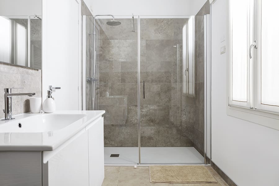 Modern bathroom. Sink with soap and shower with glass doors. Furnished with marble and beige ceramic tiles. Bright bathroom illuminated by light entering through the window